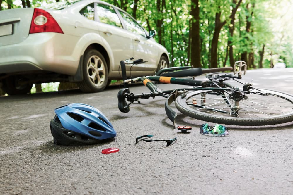 A hazardous scene unfolds on a forested road during the daytime, where a silver car has collided with a bicycle. 