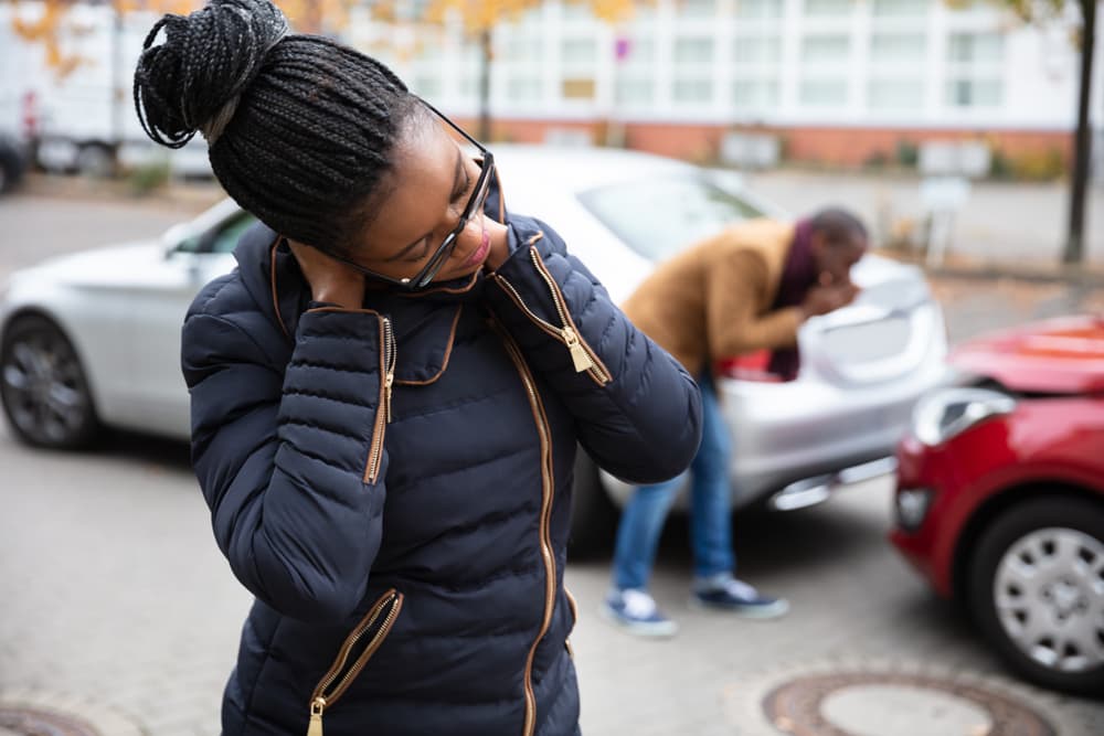 A woman experiencing neck pain is standing in front of a man as they examine a damaged car on the road.