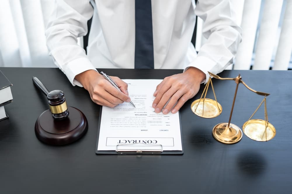 A male lawyer sits at a table in a courtroom, reviewing contract papers with law books and a wooden gavel beside him. 