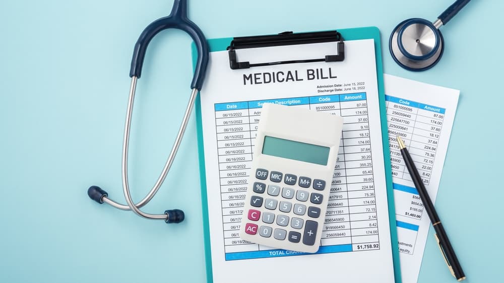 A calculator placed on top of a medical bill, accompanied by a stethoscope and a pen, all arranged on a light blue background with ample copy space.